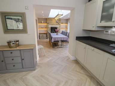 A dining room with a long table and chairs connected to a kitchen with cabinets at 2 Summers Way in Moreton-In-Marsh
