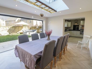 A dining room with eight chairs around a table with a cloth and flowers with a kitchen and garden visible at 2 Summers Way in Moreton-In-Marsh