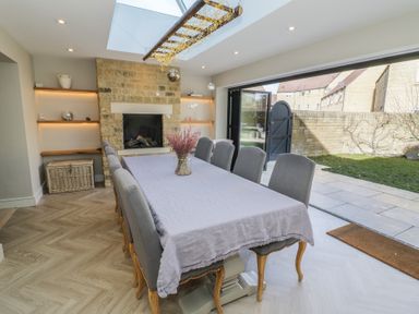 A dining room with a long table covered with a cloth surrounded by chairs a stone fireplace and glass doors leading to a garden at 2 Summers Way in Moreton-In-Marsh