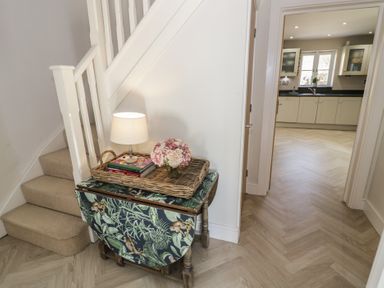 A hallway with stairs carpeted in beige and a small table holding a lamp, books, and flowers next to an open doorway leading to a kitchen with cabinets and a window at 2 Summers Way in Moreton-In-Marsh