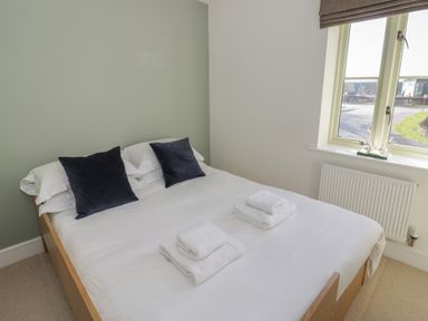 A bedroom with a bed made with white linens two black pillows and folded towels near a window at 2 Summers Way in Moreton-In-Marsh