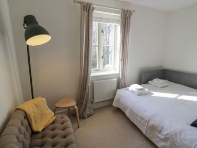 A bedroom with a bed, window with curtains, small table, sofa with a yellow blanket, and a floor lamp at 2 Summers Way in Moreton-In-Marsh