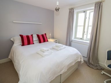 A bedroom with a double bed white bedding two red striped pillows bedside table with lamp and window with curtains at 2 Summers Way Moreton-In-Marsh