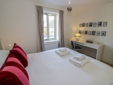 A bedroom with a bed with red and pink pillows towels on bed a window with curtains and a desk with a lamp and chair at 2 Summers Way in Moreton-In-Marsh