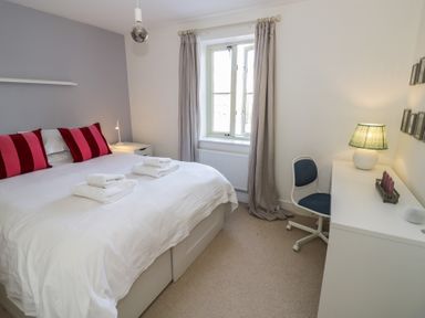 A bedroom with a white bed and red pillows next to a window and a desk with a chair and lamp at 2 Summers Way in Moreton-In-Marsh