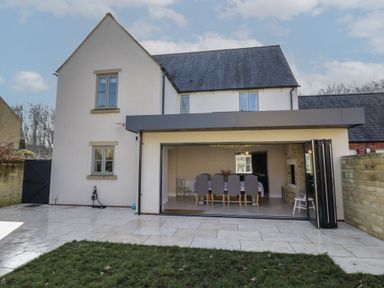 A house with a patio area and grass lawn in front with dining table and chairs visible inside at 2 Summers Way in Moreton-In-Marsh