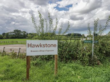 A sign for Hawkstone Brewery Shop Taproom in a grassy rural area with directional footpath signs at 2 Summers Way in Moreton-In-Marsh