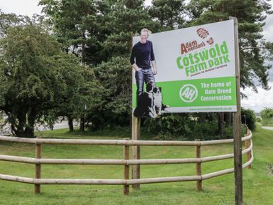 An outdoor sign for Adam Henson's Cotswold Farm Park with a man and two dogs next to wooden fencing and trees in the background at 2 Summers Way in Moreton-In-Marsh
