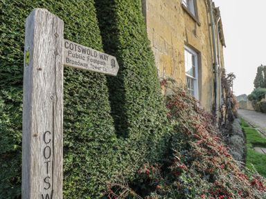 A wooden signpost for Cotswold Way public footpath near a stone building with green hedges and red berries at 2 Summers Way Moreton-In-Marsh