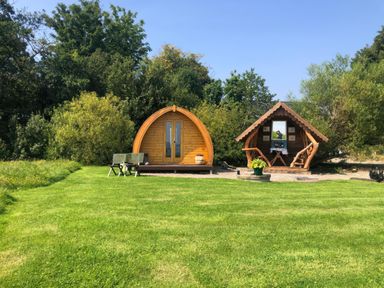 A grassy area with two wooden pods and trees in the background at River View Pod near Muff County Donegal