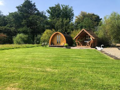 A grassy backyard with a small wooden pod and a covered wooden seating area near trees at River View Pod near Muff County Donegal