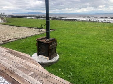 An outdoor wood stove on a grassy area near a wooden deck with a view of a rocky shoreline and distant hills at River View Pod near Muff County Donegal