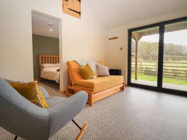 A living room with a gray rocking chair and orange futon sofa near sliding glass doors showing outdoor view at Beech Tree Cabin in Goodmanham near Market Weighton