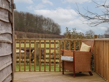 A wooden deck with a wicker chair and cushion overlooking a fenced field with trees at Beech Tree Cabin Goodmanham near Market Weighton