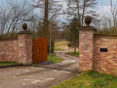 A brick entrance gate with one wooden door open a cattle grid and a winding dirt road at Beech Tree Cabin in Goodmanham near Market Weighton