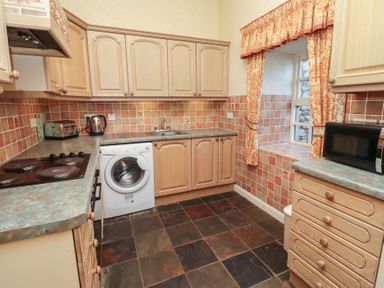 A kitchen with wooden cabinets and tiled walls featuring a washing machine toaster microwave and window with curtains at Aubretia Trail Akeld near Wooler