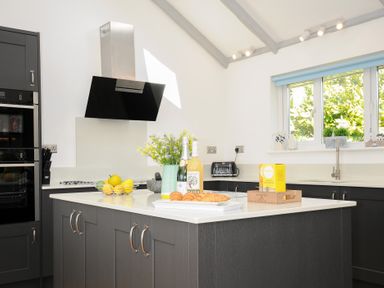 A kitchen with a black hob, a black oven, a white countertop island with croissants, bottles, flowers, a fruit bowl, and cabinets at Atlanta near Tintagel