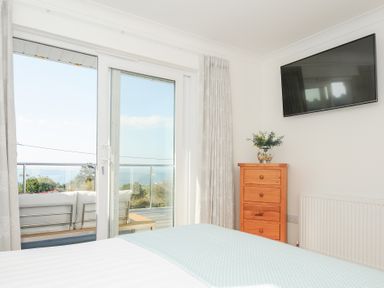 A bedroom with a bed covered with a light blue blanket a wooden chest of drawers with a plant on top a wall-mounted tv and a glass door leading to a balcony with outdoor seating at Atlanta near Tintagel