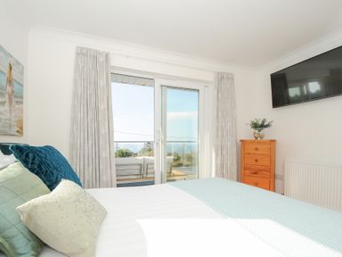 A bedroom with a bed with cushions a wooden chest of drawers a wall-mounted TV glass doors with curtains leading to a balcony with outdoor furniture at Atlanta near Tintagel