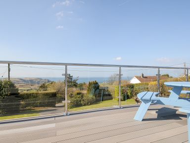 A balcony with a glass railing and a blue wooden picnic table overlooking a garden and distant hills near Tintagel