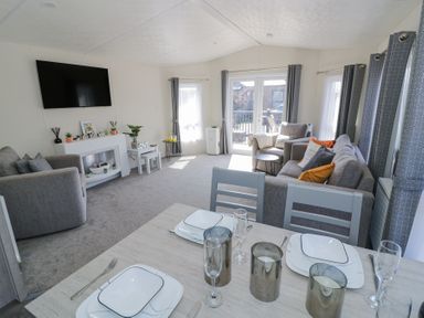 A living room with grey sofas a television on the wall and a dining table set with plates at Casa De Rhu in Evesham