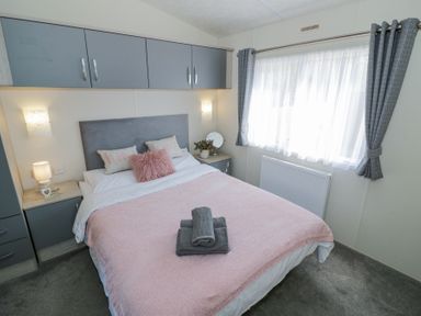 A bedroom with a bed covered in a pink blanket and gray towels on top with gray curtains and overhead cabinets at Casa De Rhu in Evesham