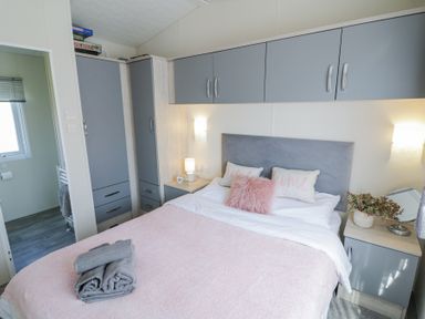 A bedroom with a bed with pink and white bedding gray towels on the bed gray wardrobe cabinets and a bedside table with a lamp and decor at Casa De Rhu in Evesham
