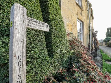 A wooden Cotswold Way public footpath sign near a green hedge and a stone building with windows at Casa De Rhu in Evesham