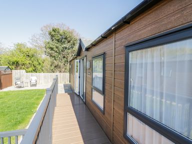 A wooden exterior walkway with railing leading to a patio and lawn with chairs at Casa De Rhu in Evesham