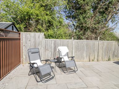Two reclining chairs with cushions and a small round table on a paved patio with a wooden fence and trees in the background at Casa De Rhu in Evesham