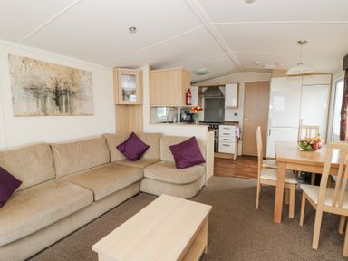 A living room with a beige sectional sofa and purple cushions a wooden coffee table a dining table with chairs and a fruit bowl and a kitchen area with cabinets and appliances at 21 Bay View in Maenygroes near New Quay Ceredigion