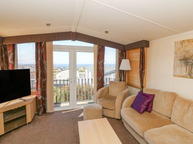 A living room with beige sofa and armchair next to glass doors with curtains and a television on a wooden stand at 21 Bay View in Maenygroes near New Quay Ceredigion
