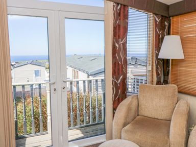 A beige armchair next to a floor lamp by windows with floral curtains and blinds overlooking a balcony with sea view at 21 Bay View in Maenygroes near New Quay Ceredigion