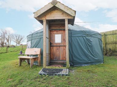 A green yurt with a wooden door and porch with a bench and small table on grass at Blaidd Bach in Carmarthen