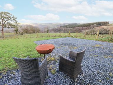 Two outdoor wicker chairs and a fire pit on a gravel area overlooking a fenced grassy field with hills in the background at Blaidd Bach in Carmarthen