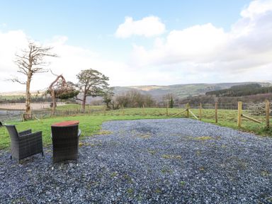 An outdoor area with two wicker chairs and a small table on a gravel surface overlooking a fenced green field with hills in the background at Blaidd Bach in Carmarthen