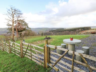 An outdoor area with a wooden fence, stone table and stools, a fire pit, and a landscape view at Blaidd Bach in Carmarthen