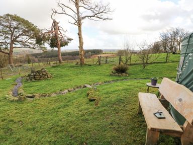 A garden with a wooden bench and small table beside a tent and a grass lawn with trees and a rock pile at Blaidd Bach in Carmarthen