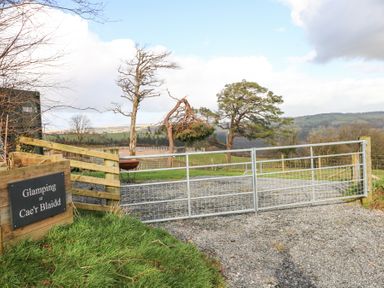 A metal gate on a gravel path with a wooden sign saying Glamping at Cae'r Blaidd and trees and hills in the background at Blaidd Bach in Carmarthen
