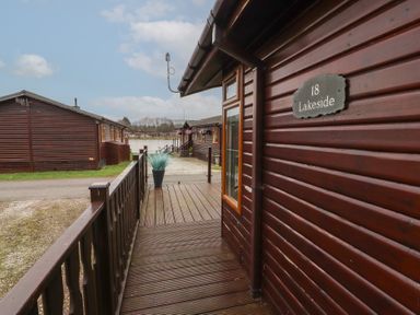 A wooden cabin with number 18 Lakeside on a sign and a walkway leading to other similar cabins near a lake at Lakeside 18 in Carnforth