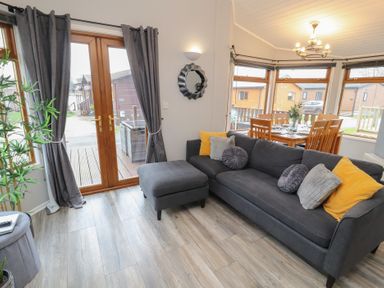 A living room with a gray sofa and ottoman near wooden doors and a dining area with wooden chairs and a table at Lakeside 18 in Carnforth