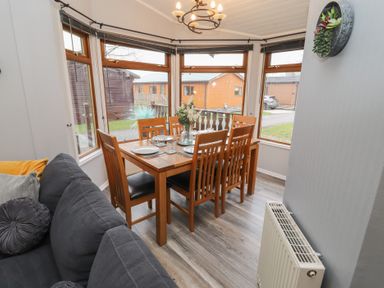 A dining area with a wooden table set for six near large windows and a gray sofa at Lakeside 18 in Carnforth