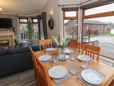 A dining table set with white plates and glasses next to large windows showing outside wooden lodges at Lakeside 18 in Carnforth