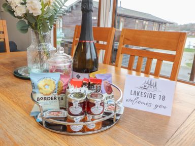 A wooden dining table with a tray of snacks jars of jam a bottle and a vase with flowers at Lakeside 18 in Carnforth