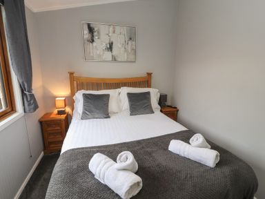A bedroom with a wooden bed frame white bedding grey pillows rolled white towels bedside tables with lamps and a window with grey curtains at Lakeside 18 in Carnforth