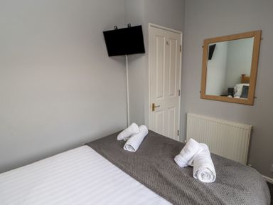 A bedroom with a bed covered with white and gray linens and rolled white towels a wall-mounted tv a door a mirror and a radiator at Lakeside 18 in Carnforth