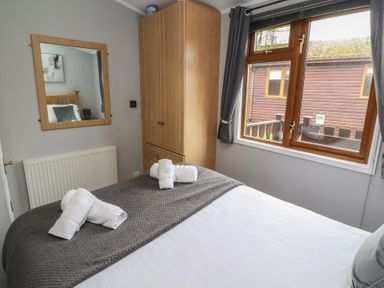 A bedroom with a bed with folded towels a wooden wardrobe a window with curtains and a wall mirror at Lakeside 18 in Carnforth