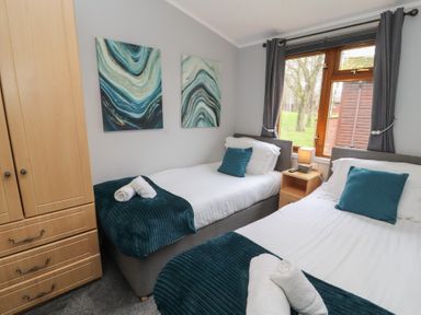 A bedroom with two single beds blue cushions and blankets a wooden wardrobe and a window at Lakeside 18 in Carnforth