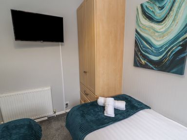 A bedroom with a bed covered with a dark teal blanket towels on the bed a wooden wardrobe a wall-mounted TV and wall art at Lakeside 18 in Carnforth