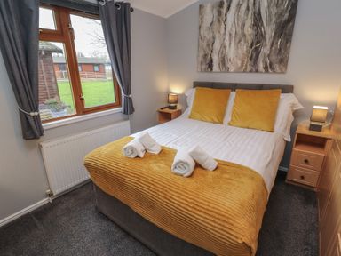 A bedroom with a double bed yellow cushions and throw two bedside tables with lamps and a window with curtains at Lakeside 18 in Carnforth